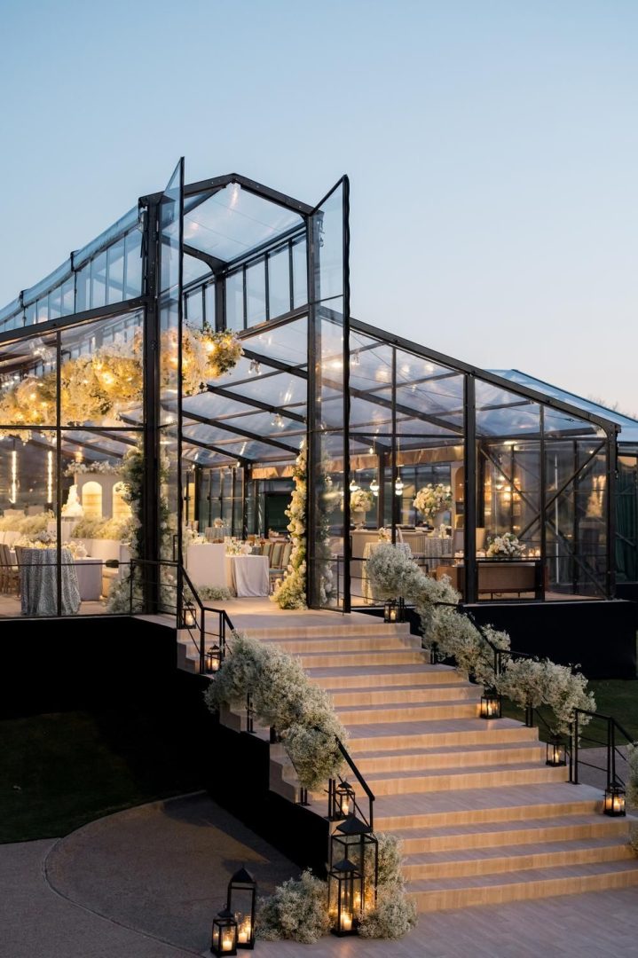 Wedding reception inside a glass venue at night, lit with ambient lighting and a romantic atmosphere.